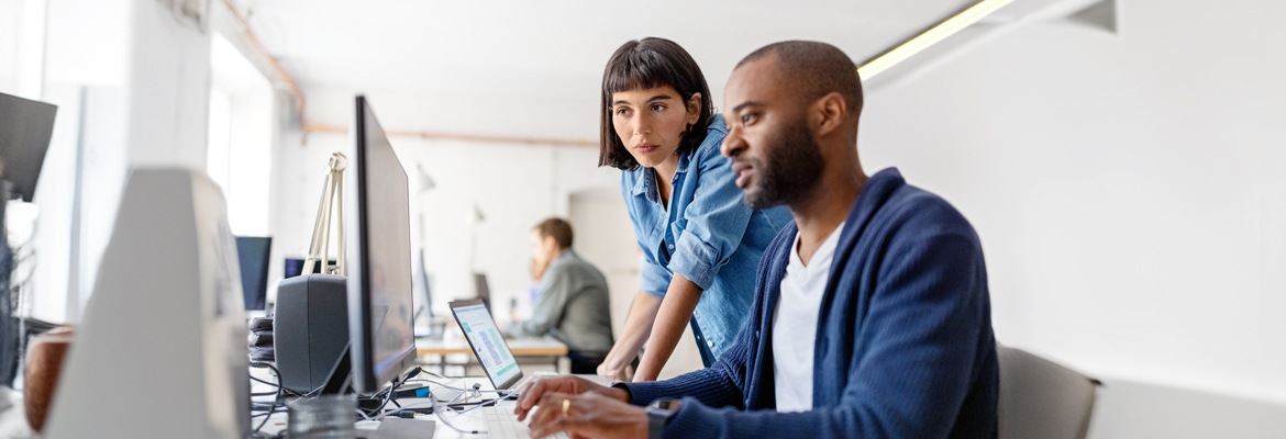 a woman looks over a man's shoulder at his computer as they work out a problem